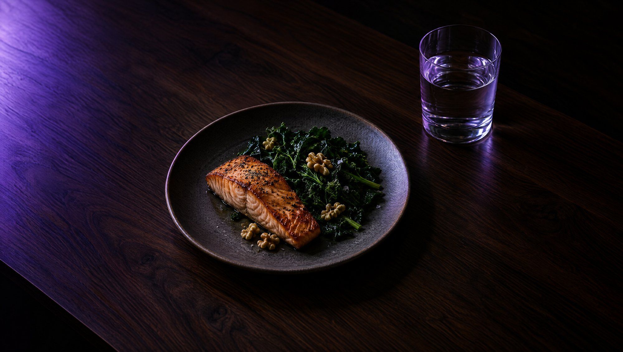 A plate of seared salmon, dark leafy greens and walnuts beside a glass of water on a dark wooden table, lit from one side in violet.